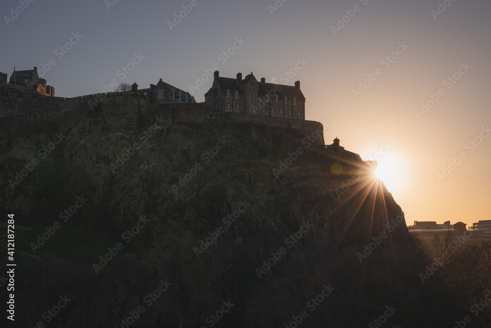 A silhouette of historic Edinburgh Castle with a sunburst at sunset or sunrise in the Scotland capital city.