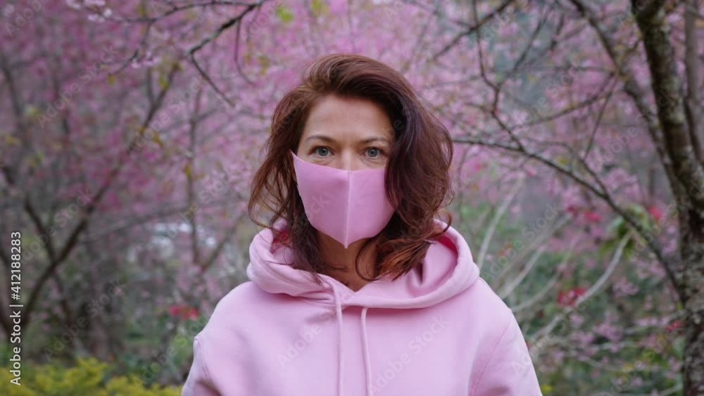 a woman in a pink hoodie and a pink protective mask stands in the sakura garden and looks directly into the camera in slow motion a woman in a pink hoodie and a pink protective mask stands in the sakura garden and looks directly into the camera in slow motion