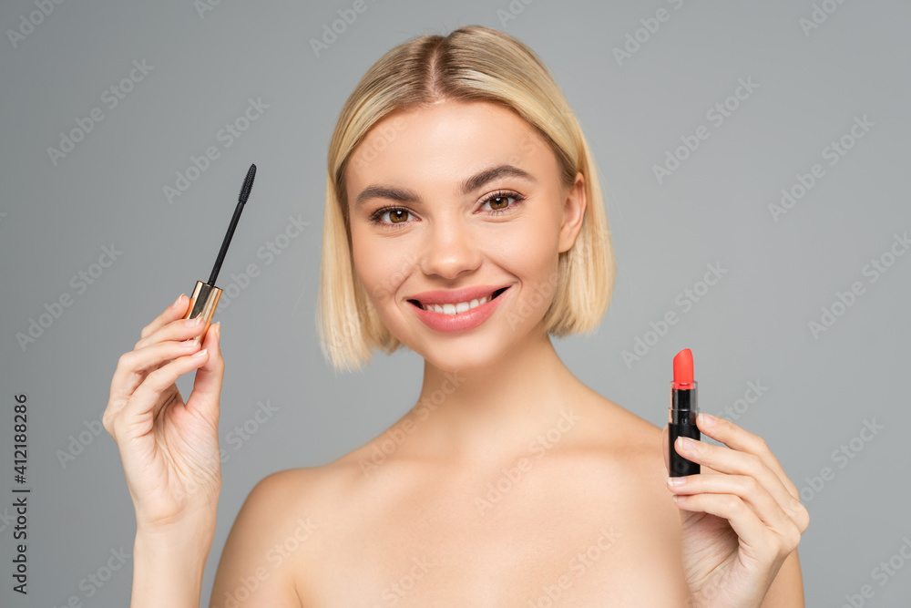 Smiling woman holding mascara applicator and red lipstick isolated on grey Smiling woman holding mascara applicator and red lipstick isolated on grey