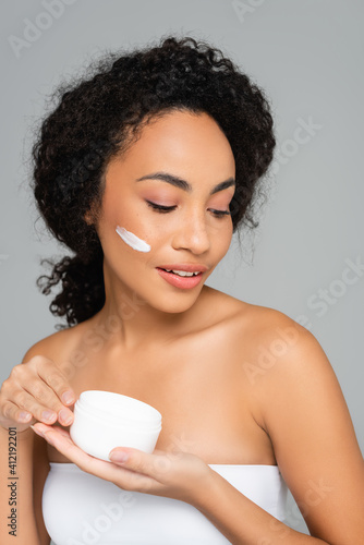 African american woman in white top holding cosmetic cream isolated on grey