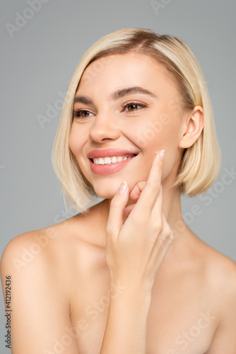 Young woman with naked shoulders applying cream on face isolated on grey