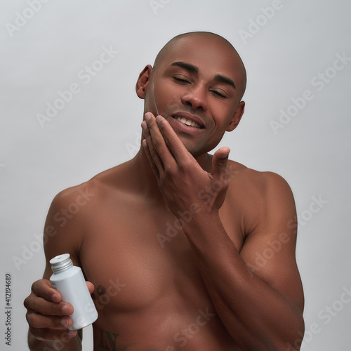 Shirtless handsome african american guy smiling, applying aftershave lotion while posing with eyes closed isolated over gray background photo
