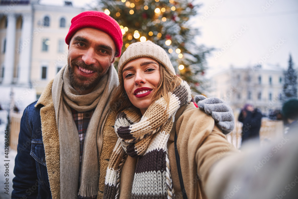 Jolly young couple taking selfie at Christmas Tree outdoors