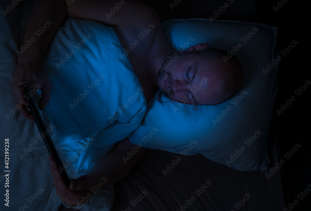 Man lying in bed at night looking at his tablet with harmful blue light illuminating his face.