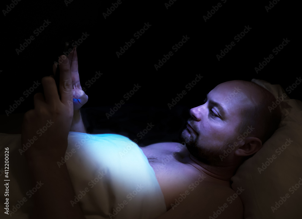 Naklejka premium Man lying in bed at night looking at his tablet with harmful blue light illuminating his face.