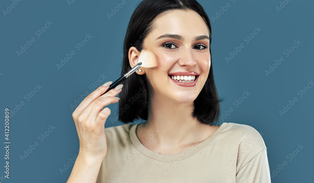 Candid portrait of a happy brunette young woman looking to the camera and making makeup with a brush herself. Candid portrait of a happy brunette young woman looking to the camera and making makeup with a brush herself.