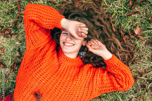 Canvas Print Overhead image of a joyful young woman wearing a knitted orange sweater lying on the grass
