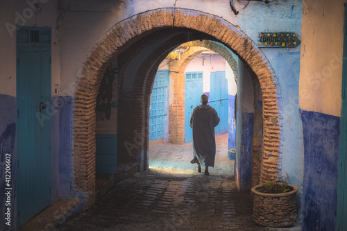A Moroccoan man in a traditional djellaba walks under an arch in a colourful street scene in the old town medina of Chefchaouen, Morocco with its blue buildings.