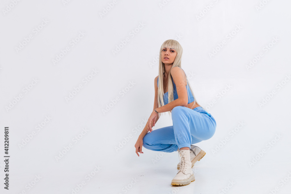 Naklejka premium Fashion portrait of young happy blonde woman with long gorgeous straight hair and bangs in the studio on a white background. The girl is dressed in a trendy blue suit, warm pants and a top