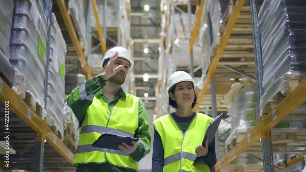 Young woman and man are talking and walking at warehouse during working day. Front view of american employees discussing work and going in storehouse, guy holding tablet in hand. People wearing