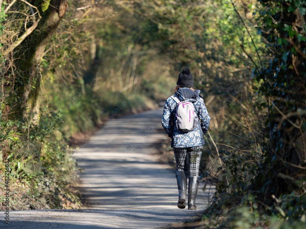 A lady walker walks down a country lane with rucksack and boots.The road is enclosed by trees and their shadows are dappled on the roadway.