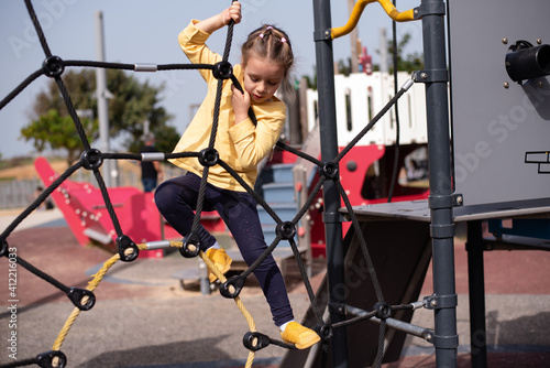 Little cute girl is playing on playground. Children's attraction from cobweb. Little girl has risen high and is holding onto ropes. Serious face. Great vacation with children.