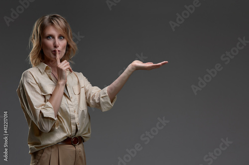 Young woman holding finger on lips mouth to keep it quiet hush pointing at copy space, standing isolated on grey blank studio background with copy space, millennial gossip girl showing shh gesture