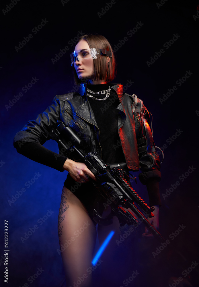 Fashionable woman with stylish haircut in black clothing poses holding a  futuristic rifle. Beautiful female soldier in cyberpunk style. Stock Photo