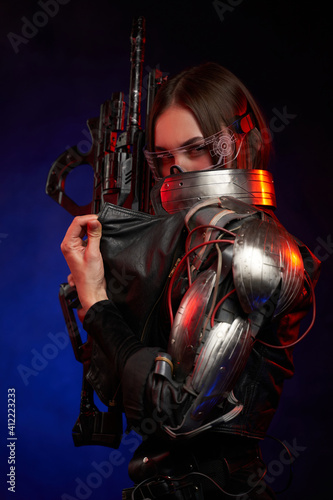 Beautiful cyber woman holding rifle and hiding her face in dark background. Dressed in black jacket martial woman in cyberpunk style looks at camera.