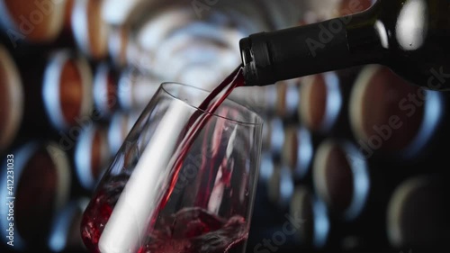 Red wine pours from the neck of the bottle into a wineglass. Oak barrels for wine in the cellar of the winery in background.