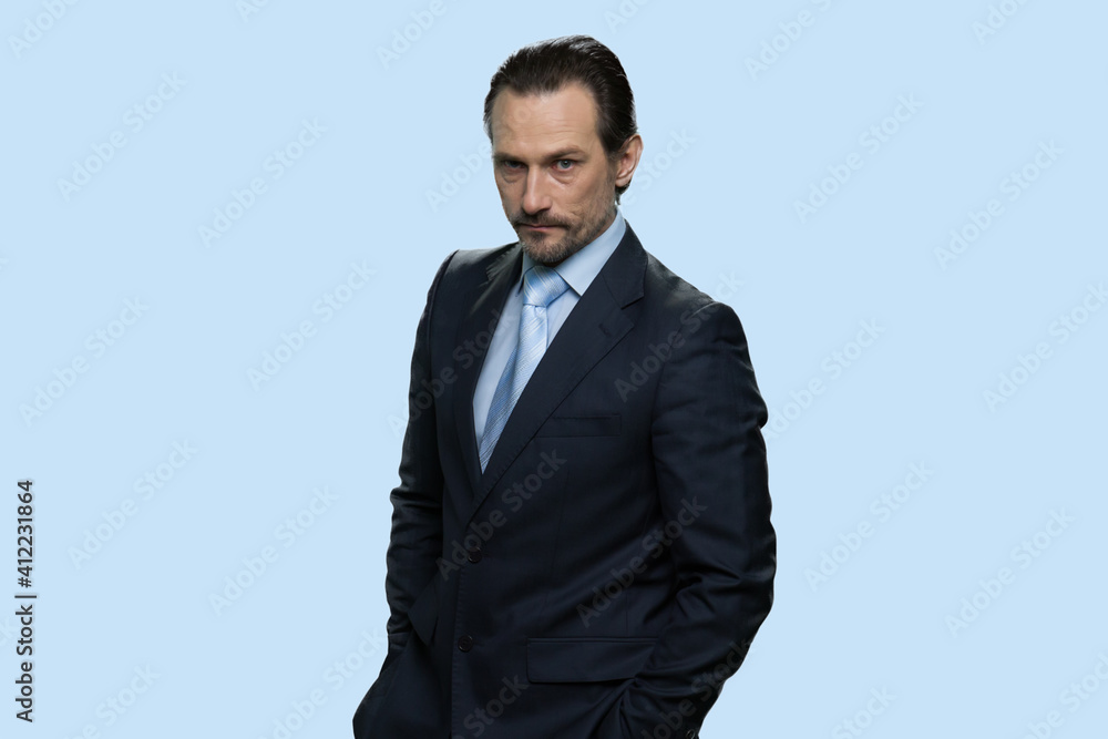 Portrait of confident mature mafia boss. Middle-aged businessman wearing protective suit and hiding his hands in pockets. Isolated on blue background. Portrait of confident mature mafia boss. Middle-aged businessman wearing protective suit and hiding his hands in pockets. Isolated on blue background.