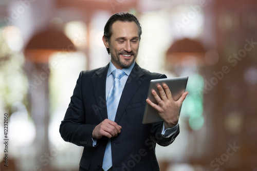 Smiling mature businessman is looking at tablet screen. Indoors restaurant on the background.