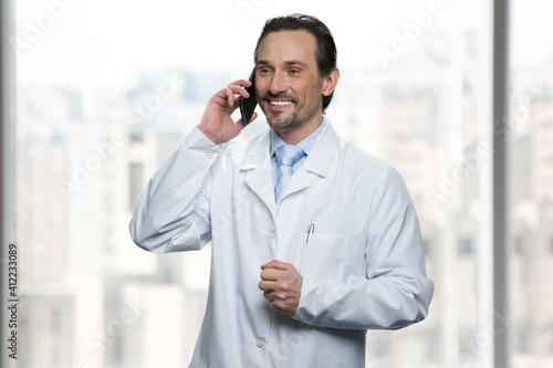 Smiling doctor is talking on phone. Bright windows on the background.