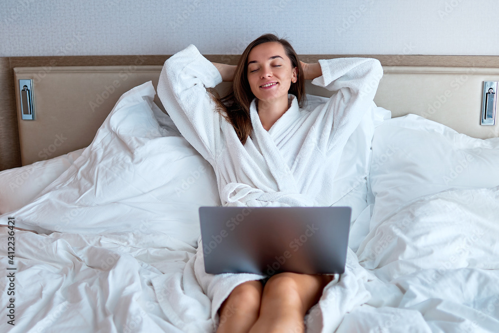 Calm smiling dreaming woman freelancer with hands behind head wearing white bathrobe remote working online at a computer on the bed from a hotel room. Easy lifestyle and satisfaction Calm smiling dreaming woman freelancer with hands behind head wearing white bathrobe remote working online at a computer on the bed from a hotel room. Easy lifestyle and satisfaction