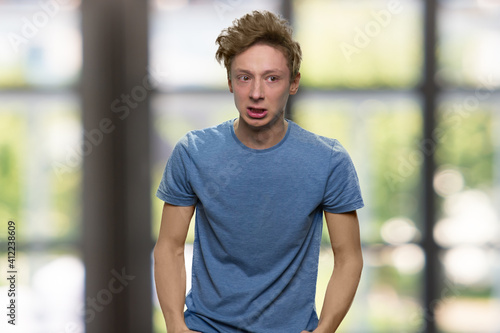 Sad teenager against the window. Unhappy american boy in t-shirt. Blurred window on the background.
