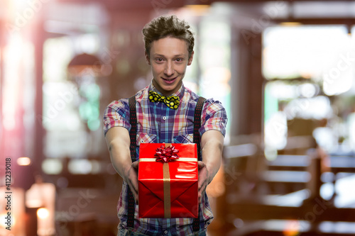 Handsome young boy is giving you a present. Schoolboy in checkered t-shirt with bowtie and black suspenders. Blurred indoors on the background. photo