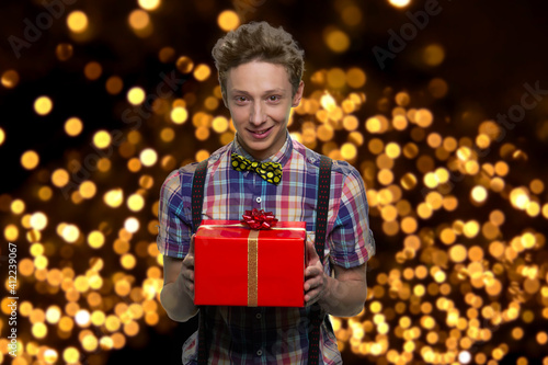 Portrait of smiling welll-dressed boy with red gift box. Handsome guy is standing against background with glowing lights. photo