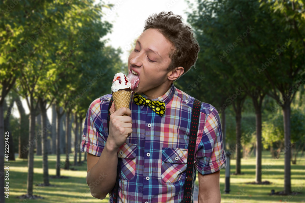 Fototapeta premium American teen boy in suspenders is licking ice-cream. Green park on the background.