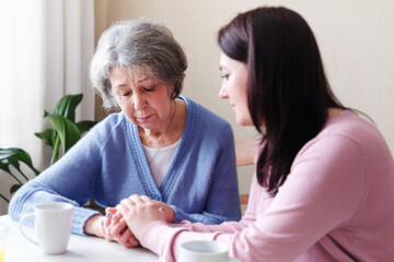 Sad elderly woman is comforted by a young woman and holds her hand - Girl volunteer supports a pensioner - The concept of family relationships and mental health