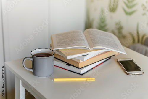 A stack of books, a mug of tea, and a mobile phone on a white table. A bookcase with books in the background in a modern interior. The concept of training and education.