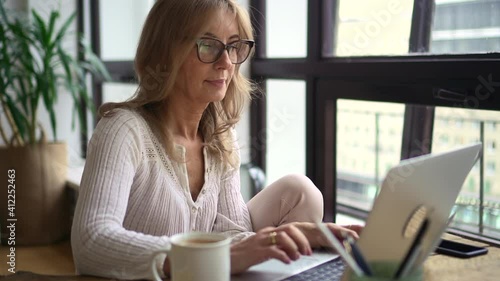 Businesswoman working with laptop computer and sitting at table in home office. Middle aged American woman looks at screen of device with wireless and does remote work, sits at desk in light room