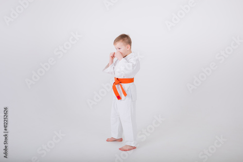 small boy in a white kimono with an orange belt stands sideways in a protective pose against a white background with a copy of the space