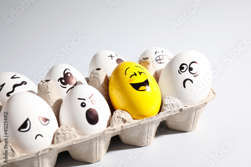 Yellow smiley egg among others with negative emotions in package on light background photo