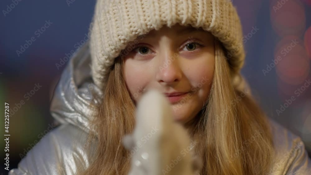 Close-up of beautiful Caucasian girl rubbing hands in mittens outdoors in the evening. Headshot of cute smiling child enjoying winter weather at dusk with garlands at background. Christmas time. Close-up of beautiful Caucasian girl rubbing hands in mittens outdoors in the evening. Headshot of cute smiling child enjoying winter weather at dusk with garlands at background. Christmas time.