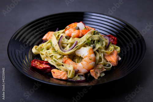 Noodles with seafood, sun-dried tomatoes, capers and red onions. Homemade pasta with shrimp, salmon (trout) and pesto sauce. Black background, black plate. Close-up.