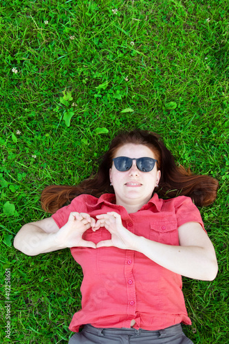Relaxed woman lying on the grass. Young caucasian girl in sunglasses shows love heart sign with her hands