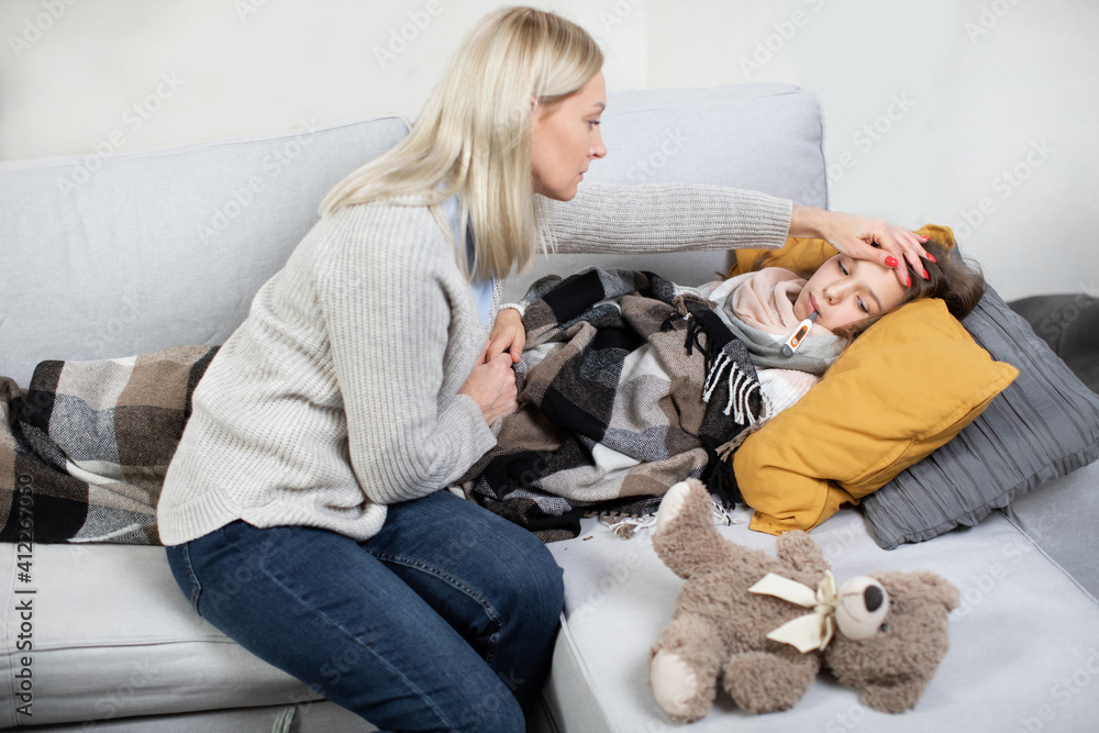 Flu, cold, and fever concept. Good-looking confident caring middle aged mother touching forehead of her sick sad daughter, lying in bed with fever and thermometer in the mouth Flu, cold, and fever concept. Good-looking confident caring middle aged mother touching forehead of her sick sad daughter, lying in bed with fever and thermometer in the mouth