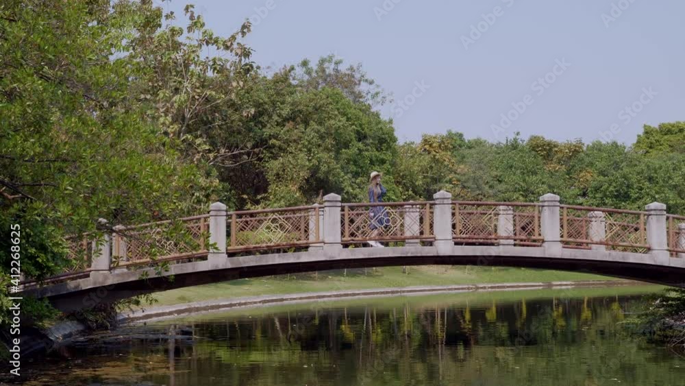 Happy Travel Woman on Holiday Walking and Relax on Bridge Over River in City Park with Green Trees. Beautiful Female Traveller on Vacation in Tropical Forest in Hot Summer Day in Thailand Happy Travel Woman on Holiday Walking and Relax on Bridge Over River in City Park with Green Trees. Beautiful Female Traveller on Vacation in Tropical Forest in Hot Summer Day in Thailand