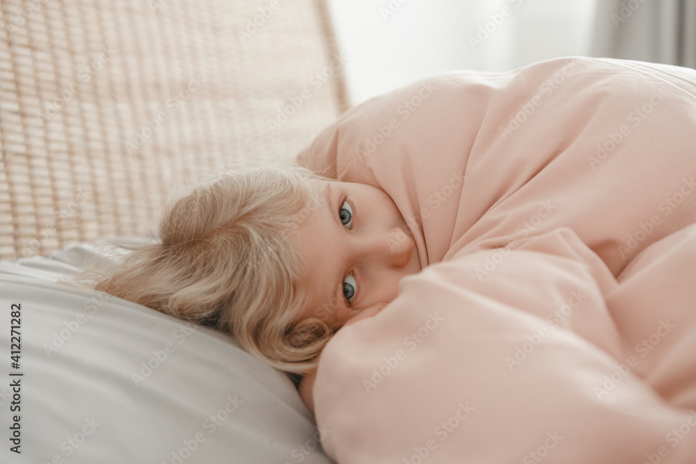 Pensive Caucasian blonde child girl lying on pillow under blanket in bed at home. Sad kid in stress or frustration. Child lying with open eyes thinking or dreaming. Candid authentic home life. Pensive Caucasian blonde child girl lying on pillow under blanket in bed at home. Sad kid in stress or frustration. Child lying with open eyes thinking or dreaming. Candid authentic home life.