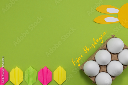 Dutch words Fijne Paasdagen (happy Easter) on a green background with yellow paper bunny ears and paper tulips. White eggs in a cardboard carton. 