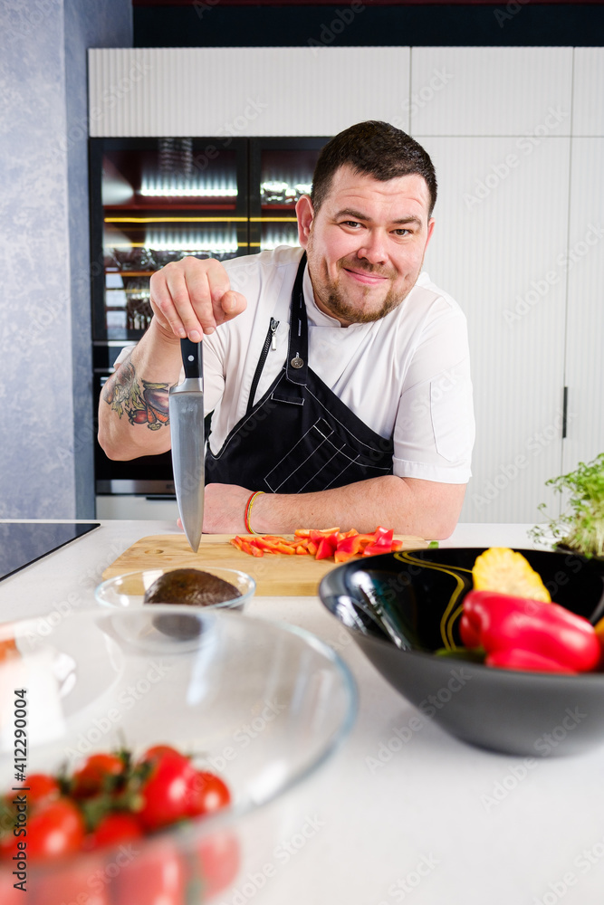 40 years man chef cooker in a white uniform and a black apron in the kitchen of the restaurant prepares a salad of vegetables and fish. 40 years man chef cooker in a white uniform and a black apron in the kitchen of the restaurant prepares a salad of vegetables and fish.