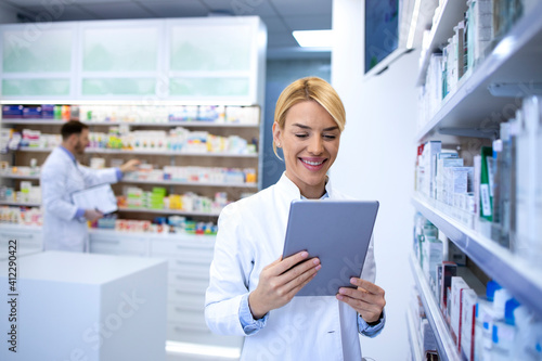 Portrait of female experienced pharmacist working on tablet in pharmacy store by the shelf with medicines.