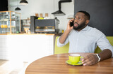 Puzzled African-American guy is sitting in the cafe with cup of coffee and has serious phone conversation, a multiracial man talking on the smartphone