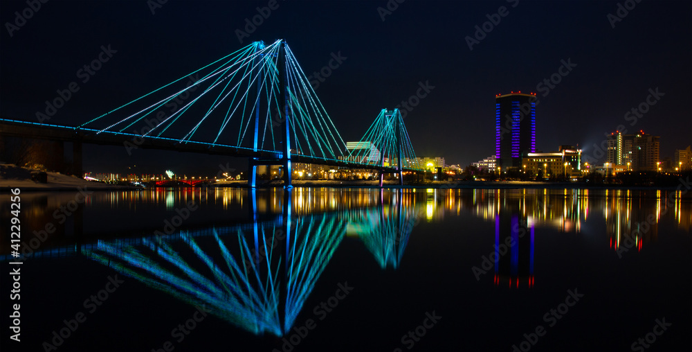 Night landscape with a bridge over the Yenisei River in Krasnoyarsk, Russia. Blurred reflection bright lights in the dark water.