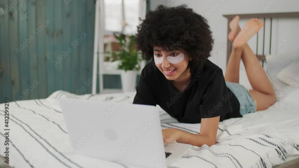 Skin Care, Cosmetics at Home, Online Learning, Remote Work, Afro American Freelancer. Afro american woman with eye patches lying on bed using laptop Skin Care, Cosmetics at Home, Online Learning, Remote Work, Afro American Freelancer. Afro american woman with eye patches lying on bed using laptop