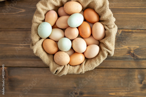 A group of organic farm fresh eggs in a burlap cloth bowl sitting on a dark wood table. Collection includes dark brown eggs, tan eggs and blue eggs. 