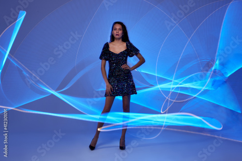 female in fashionable wear posing at camera against the background of freeze light lines, young woman is confidently looking, abstract lines drawn by light