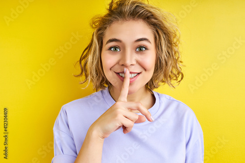 smiling woman asking to be quiet with finger on lips. silence and secret concept. curly female looks at camera, close-up portrait