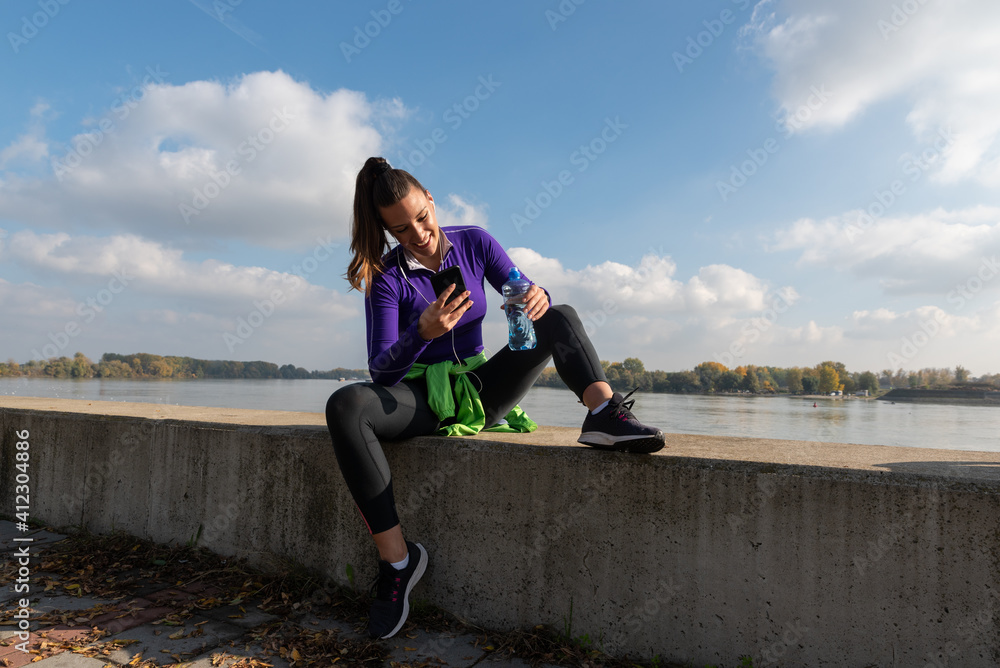 Young sweaty active fitness athletic woman taking a break after outdoor exercising of running and jogging changing the music on her smartphone Young sweaty active fitness athletic woman taking a break after outdoor exercising of running and jogging changing the music on her smartphone