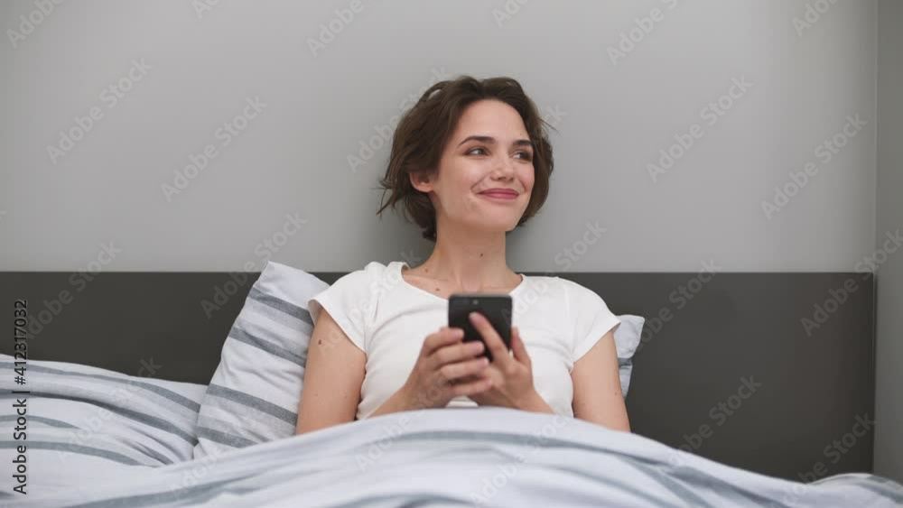 Calm young woman wears white t-shirt lying in bed rest relax spend time in bedroom lounge home in own room house hotel hold in hands using mobile cell phone chatting order delivery Real estate concept Calm young woman wears white t-shirt lying in bed rest relax spend time in bedroom lounge home in own room house hotel hold in hands using mobile cell phone chatting order delivery Real estate concept